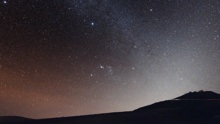 A photograph of space from the ground. On the bottom of the image is land, which leads to a mountain on the right. Above the land is space, shown in orange, black and white.